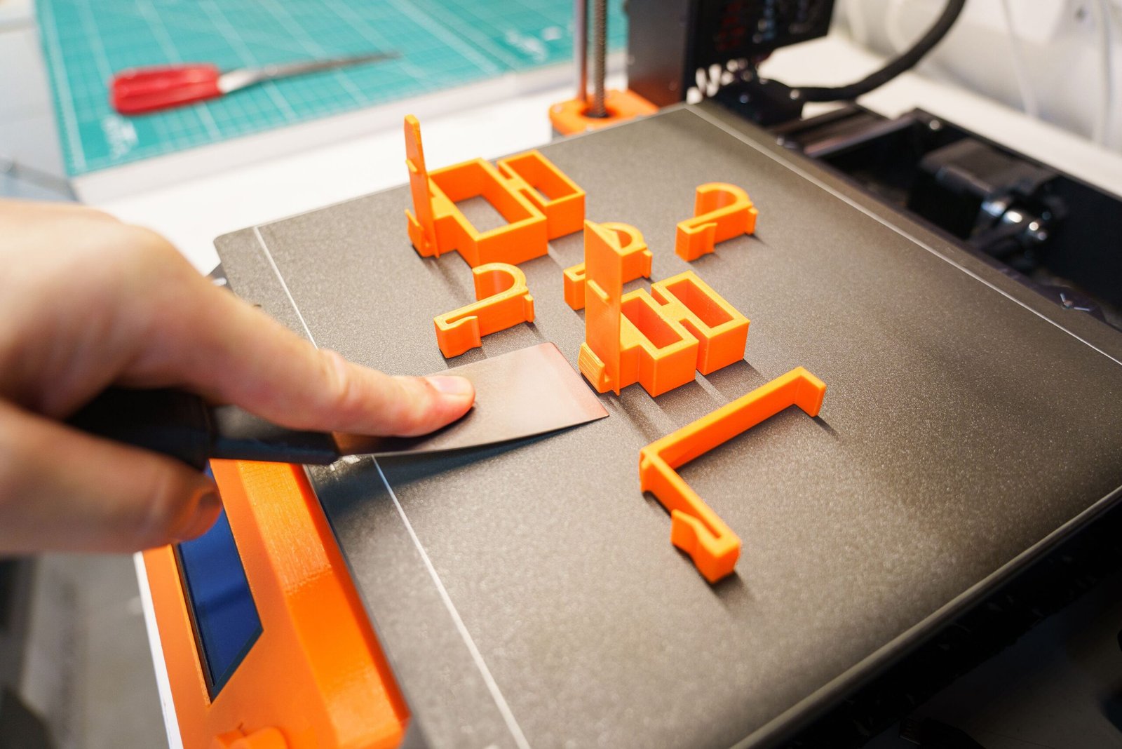 Home A hand carefully removes an orange 3D printed object from a printer bed using a spatula indoors.