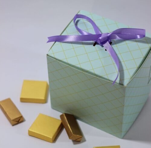 Home A stylish gift box tied with a purple ribbon, surrounded by gold-wrapped chocolates, perfect for celebrations.